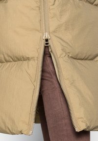 Chaqueta acolchada beige con una superficie brillante y texturizada, que cuenta con una cremallera de longitud completa y un diseño acolchado. Tela marrón visible debajo.