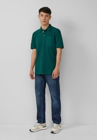 Zielona koszulka polo z krótkim rękawem i kieszonką, zestawiona z ciemnoniebieskimi dżinsami oraz szaro-białymi trampkami z zielonymi akcentami. Prosty design.