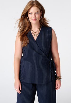 Femme aux longs cheveux roux portant un top cache-cœur sans manches bleu marine à rayures fines et un pantalon assorti, accessoirisée avec des colliers et bracelets superposés.