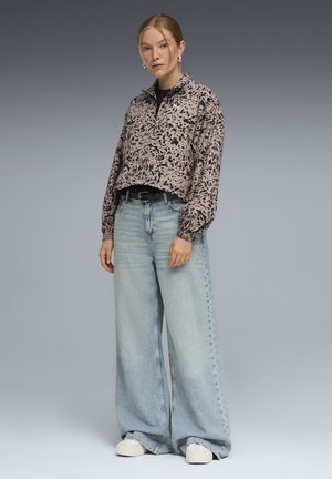 Γυναίκα που φοράει άνετο πουλόβερ με λεοπάρ σχέδιο, ανοιχτόχρωμα τζιν με φαρδιά μπατζάκια, λευκά αθλητικά παπούτσια και ασημένια κρίκους σκουλαρίκια, μπροστά σε γκρι φόντο.