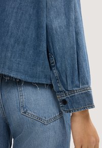 Close-up of a person wearing a blue denim shirt with buttoned cuffs and matching blue denim jeans with a visible back pocket.