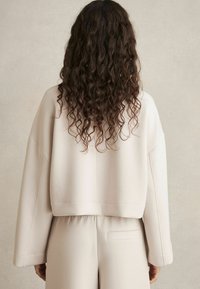 Felpa cropped beige con maniche ampie, realizzata in tessuto morbido. Presenta un dettaglio di cucitura ed è abbinata a pantaloni beige a vita alta.