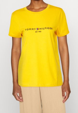 Osoba nosząca jaskrawożółtą koszulkę Tommy Hilfiger z logo i napisem "Est. 1985", w połączeniu z jasnobeżowymi spodniami.