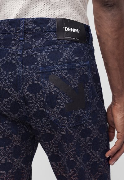 Jean en denim bleu foncé à motifs avec logo flèche noir sur la poche arrière et étiquette noire indiquant "DENIM" sur la ceinture, porté par une personne.