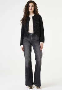 Zwarte tweed jas met subtiele textuur, ronde halslijn en twee knopen, gecombineerd met grijze uitlopende jeans en metallic hakken.