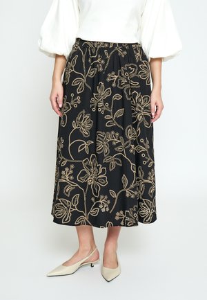 Nainen pukeutuneena mustaan midihameeseen, jossa on beigejä kukkaistikkausta, valkoinen pussihiha-paita ja beiget, kärjestä terävät slingback-korkokengät.