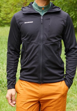 Veste softshell noire avec une coupe ajustée, poches zippées et capuche. Présente des accents orange contrastants et une couche intérieure verte.