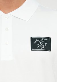 Tricou polo alb cu un guler cu trei nasturi și o pată rectangulară neagră cu text alb scris de mână. Textul spune "din Paris, iubire, Karl."