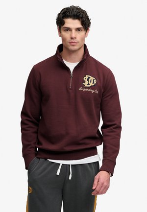 Young man wearing a maroon quarter-zip sweatshirt and gray sweatpants with yellow stripes, standing with hands in pockets.