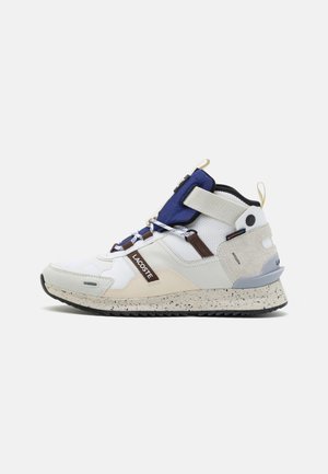 Sneakers Lacoste z białą cholewką, granatowymi akcentami, teksturowaną skórą i nakrapianą podeszwą. Oferują elastyczne sznurowadła i uchwyt z tyłu dla łatwego zakupu.