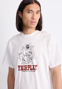 Witte katoenen t-shirt met cartoonafbeeldingen van twee personages die sport spelen, met "FAIRPLAY" in rode tekst en "CARHARTT WIP 1989" daaronder.