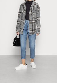 Žena v šedobéžovém kostkovaném saku, černém topu, uvázaných modrých džínách, bílých teniskách a s černou kabelkou na pozadí bez vzoru.