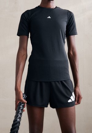 Person trägt ein schwarzes Sportshirt und Shorts mit weißen Logos und hält ein dickes schwarzes Kampfseil in der rechten Hand.