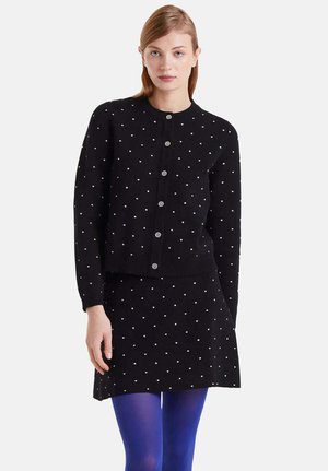 Cárdigan negro de punto con cierre de botones, adornado con pequeños lunares blancos, combinado con una falda negra a juego. Incluye medias azules.