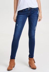 Jeans ajustados azules de denim, con parches desgastados. Llevados con una camiseta blanca casual y botas ankle marrones.