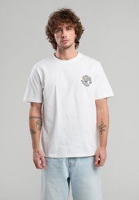 Camiseta de algodón blanca con un estampado gráfico en negro y amarillo en el lado izquierdo del pecho. Diseño de cuello redondo con mangas cortas y ajuste relajado.