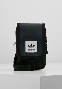 Borsa a spalla in tessuto nero con pattina ripiegabile, chiusura con zip e una targhetta del logo Adidas bianco. Dotata di una tracolla regolabile. Design compatto.