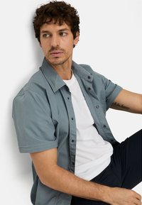 Grå kortærmet skjorte med knappelukning foran, båret over en hvid T-shirt. Glat stof med en afslappet pasform, med en brystlomme.