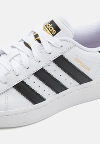 Vit lädersneaker med svart tre-randig design, texturerade accenter och guldbranding. Har vita snören och en gummisula.