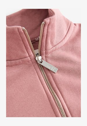 Jachetă roz din fleece cu fermoar, prevăzută cu guler înalt, fermoar metalic argintiu cu etichetă și textură moale. Design minimalist, fără modele sau accente.