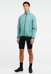 Lengvas, mėtų žalios spalvos dviratininko švarkas su aukštu apykakle ir juoda užtrauktuku. Nešiojamas virš juodų dviratininkų šortų ir batų, pasižymintis elegantišku dizainu.