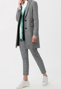 Cappotto grigio e nero a scacchi con colletto a punta, sovrapposto a una camicia a righe bianca e verde, abbinato a pantaloni a quadri grigi e sneakers bianche.