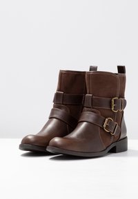 Botas de tobillo marrones que combinan cuero liso y ante suave, con correas contrastantes y hebillas en tono dorado. Tacón bajo y cuadrado.