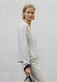 Světle šedý dlouhý rukáv s výstřihem do V a spadlými rameny, doplněný o světle šedé kalhoty. Rukávy mají černé manžety.