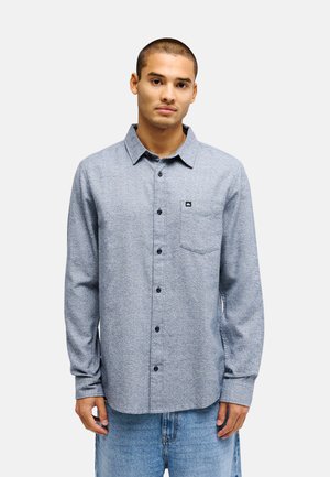 Junger Mann mit kurzen Haaren, der ein langärmliges graues Hemd mit Knöpfen und hellblaue Jeans trägt, steht vor einem schlichten Hintergrund.