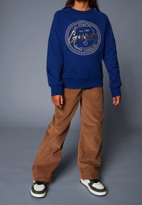 Blå bomullströja med guld-logotyp, parat med bruna kordfläskbyxor med vida ben. Svarta och gröna sneakers med vit sula.