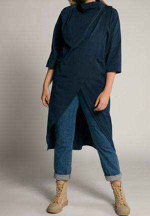 Femme portant une longue tunique asymétrique bleu foncé par-dessus un jean bleu retroussé et des bottines beiges à lacets, debout devant un fond uni.