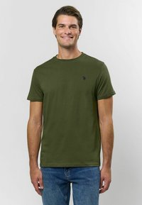 Olivově zelené bavlněné tričko s kulatým výstřihem a krátkými rukávy, s malým tmavým logem na hrudi. Nosené s modrými džínovými kalhotami.