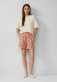 Beige übergroßes T-Shirt, kombiniert mit korallenfarbenen Shorts mit einem zarten abstrakten Leopardenmuster. Weiße Slippers runden den Look ab.