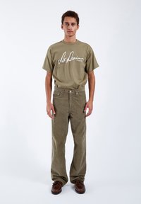 Světle olivově zelené bavlněné tričko s bílým logem, doplněné o texturované světle olivové manšestrové kalhoty. Celý outfit zakončují hnědé šněrovací boty.