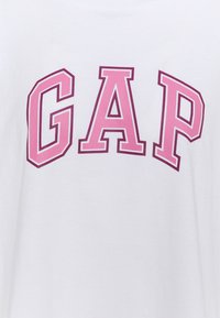 Bílý bavlněný tričko s velkým růžovým logem "GAP". Krátké rukávy, kulatý výstřih, hladká textura a mírně volný střih.