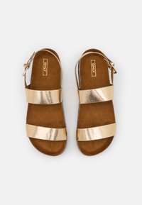 Kullanışlı ve rahat bir düz taban tasarımı ile iki geniş kayışa, ayarlanabilir bilek tokasına ve kahverengi yastıklı tabana sahip altın metalik sandaletler.