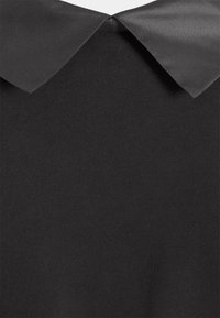 Černý pletený top s lesklým černým límcem. Materiál je hladký a přiléhavý, což kontrastuje s žebrovaným svetrkovým materiálem.