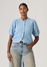 Mujer vistiendo un cárdigan corto de manga corta tejido de color azul claro con botones, combinado con jeans azul deslavado de talle alto, de pie con una mano en el bolsillo.