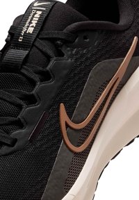 Czarny syntetyczny but sportowy z siateczkową cholewką, kontrastującym brązowym logo Nike i białą podeszwą środkową. Posiada fakturowane nałożenia i sznurowany fason.