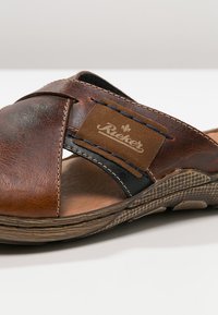 Sandal en cuir marron avec des sangles entrecroisées et un patch logo. Semelle texturée avec une combinaison de couleurs marron et noir, présentant de subtils détails de couture.