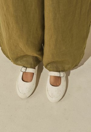 Piedi con scarpe Mary Jane bianche ricamate con cinghie a fibbia d'argento, abbinate a pantaloni larghi di lino verde oliva.