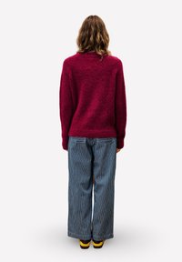 Person mit schulterlangen welligen Haaren, der einen roten Pullover, weit geschnittene blau-weiße gestreifte Hose und braune sowie gelbe Schuhe trägt, steht mit dem Rücken zur Kamera.