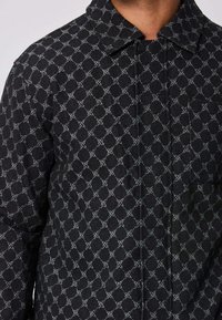 Veste noire avec un subtil motif géométrique en chaîne grise, col, fermeture à boutons-pression et poche poitrine portée par une personne recadrée au niveau du cou.