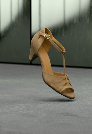 Beige mokkanahkainen T-strap-keskikorkokengäs leijuu harmaan maton yllä, taustalla heijastava tummanvihreä seinä.