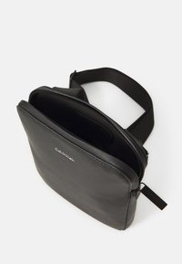 Svart läder crossbody-väska med en texturerad yta, dragkedjestängning och ett tygband. Har en logotyp på framsidan och ett internt fack.