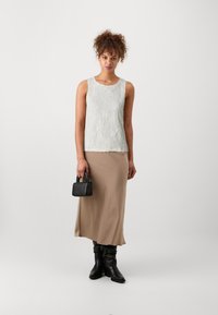 Top sin mangas texturizado de color verde claro, falda maxi de satén beige, botas negras hasta la rodilla y un pequeño bolso negro. Fondo neutro.