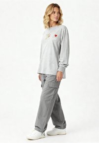 Grijze longsleeve met geborduurd ontwerp en tekst, gecombineerd met losse grijze cargobroek en witte sneakers.