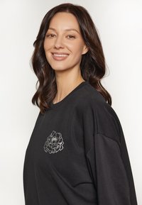 Sudadera negra con cuello redondo y corte oversize, con bordado floral blanco en el lado izquierdo del pecho. Textura suave, diseño casual.