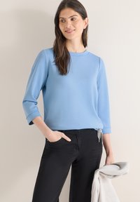 Femme aux cheveux bruns portant un haut bleu clair à manches longues et un pantalon noir, tenant un vêtement gris clair plié dans la main droite.