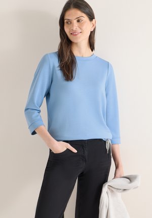 Mujer con cabello castaño que lleva una blusa de manga larga azul claro y pantalones negros, sosteniendo una prenda plegada de color gris claro en la mano derecha.